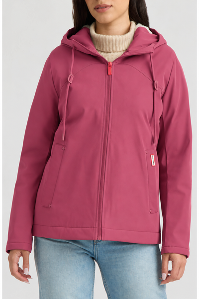 A woman wearing a dusty rose pink hooded zip-up jacket over a cream turtleneck sweater and blue jeans, shown from the nose down against a light gray studio background.